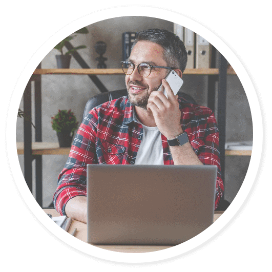Man wearing glasses and a red plaid shirt sits at a desk with a laptop, smiling while talking on a smartphone, enjoying friendly contact in his home office setting.